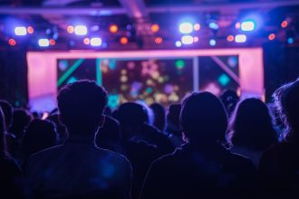 Pessoas vistas de costas acompanham um show enquanto luzes coloridas iluminam o palco.