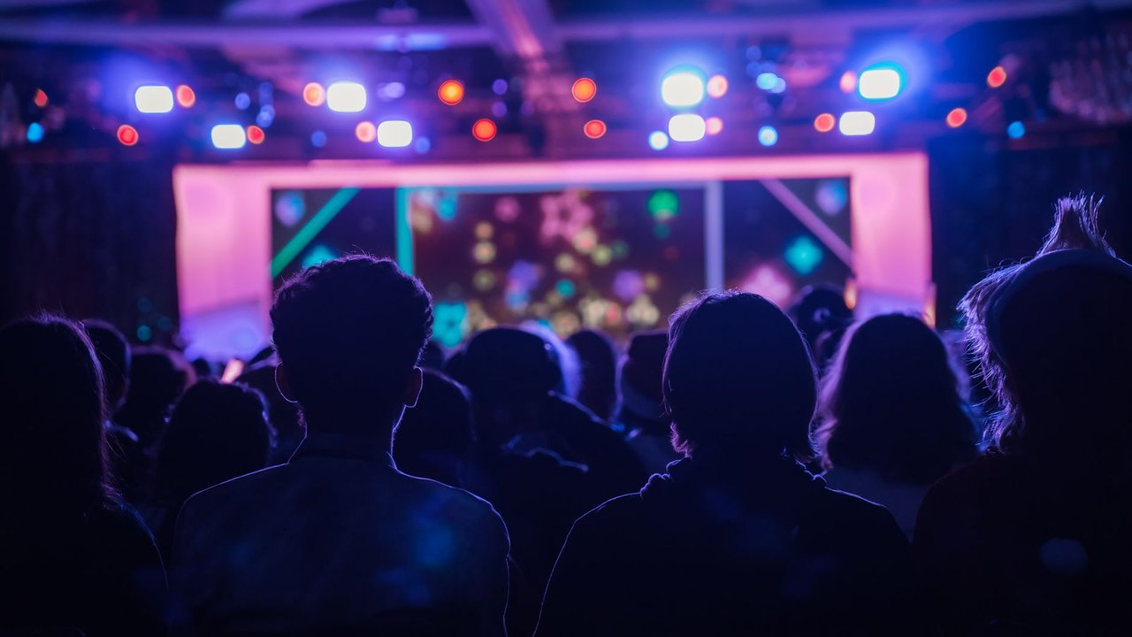 Pessoas vistas de costas acompanham um show enquanto luzes coloridas iluminam o palco.