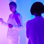 Duas pessoas interagem com interfaces digitais em um ambiente futurista iluminado por cores vibrantes.
